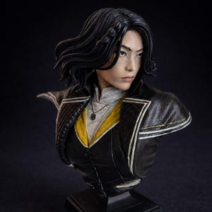 Pode incluir: Escultura de busto detalhada de uma pessoa com cabelo escuro, usando uma jaqueta preta com detalhes dourados sobre um colete amarelo. A escultura tem uma expressão séria e um colar com pingente. O fundo é escuro.
