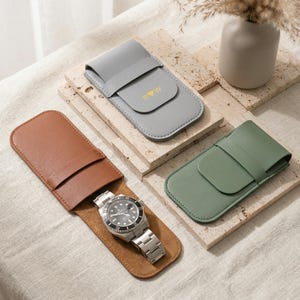 Könnte beinhalten: Drei Leder-Uhrenetuis in Braun, Grau und Grün. Das braune Etui enthält eine silberne Armbanduhr mit schwarzem Zifferblatt. Das graue Etui hat goldene Initialen. Die Etuis liegen auf einer neutralen Oberfläche mit einer Vase mit Trockenblumen.
