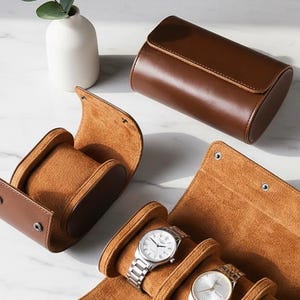 May include: A brown leather watch case with three silver watches displayed. The case is open, revealing a soft, tan interior. A separate, closed brown leather watch case and a single watch holder are also visible.