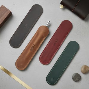 May include: Four leather pen cases in gray, brown, red, and green. The brown case holds a fountain pen. The gray case has "LN" embossed, the brown case has "CRAT", and the red case has "EST.2024". The green case has a leaf symbol.