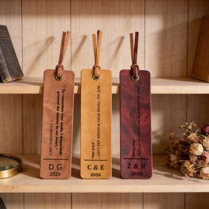 May include: Three leather bookmarks in shades of brown, tan, and burgundy, each with a leather loop. The bookmarks feature quotes and initials with the year 2026. They are displayed on a wooden shelf.