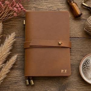 May include: A brown leather journal with a horizontal strap closure secured by a brass stud. The cover features the embossed initials P.T. in the bottom right corner. Two small brass charms hang from the bottom edge of the notebook cover.