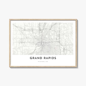 May include: A framed map print of Grand Rapids, Michigan, USA. The map is in grayscale, showing streets and waterways. The city name is printed in a bold, sans-serif font at the bottom, with the state and country below it. The frame is a light wood colour.