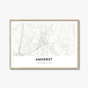 Könnte beinhalten: Ein gerahmter Kartendruck von Amherst, Massachusetts, USA. Die Karte ist in Graustufen gehalten und zeigt Straßen, Gebäude und geografische Merkmale. Der Name der Stadt ist unten zentriert, darunter "Massachusetts, USA". Der Rahmen ist hellholzfarben.
