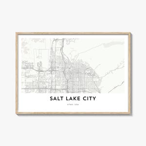 May include: A framed print showing a detailed map of Salt Lake City, Utah, in shades of grey. The city's name is in bold black text at the bottom, with "Utah, USA" beneath. The frame is a light wood colour.