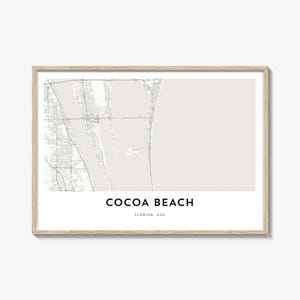 May include: A framed print featuring a detailed map of Cocoa Beach, Florida, USA. The map is in grayscale, with the city name and state printed in black at the bottom. The frame is a light wood color.