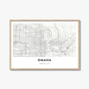 Puede incluir: Una impresión enmarcada con un mapa detallado de Omaha, Nebraska, en escala de grises. El nombre de la ciudad se muestra en la parte inferior, con "Nebraska, USA" debajo. El marco es de color madera clara.