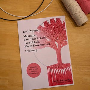Könnte beinhalten: Ein DIY-Makramee-Set mit Anleitungen für ein "Lebensbaum"-Design. Das Bild zeigt eine gedruckte Anleitung mit einer roten Baumillustration sowie Spulen mit weißem und rosa Kordel. Das Baumdesign hat einen Durchmesser von ca. 30 cm.