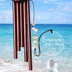 May include: A bronze wind chime with a circular top and multiple cylindrical tubes. A strand of seashells and starfish hangs below the tubes, and a clear acrylic disc with the names "Emerson Josh + Megan EST. 2024" is suspended below.