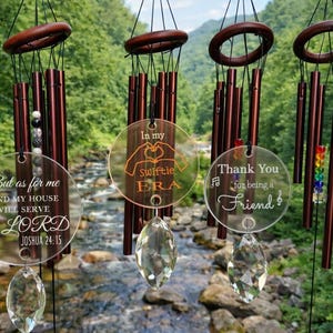 May include: Four wind chimes with brown wooden tops and tubes hang in front of a blurred natural background. Each chime has a clear, circular pendant with engraved text. One says "But as for me and my house we will serve the LORD Joshua 24:15."