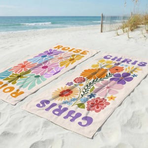 May include: Two rectangular beach towels with a cream background, each featuring a colorful floral pattern and a name printed in large, bold letters. One towel displays the name ROISE in orange, and the other displays the name CHRIS in purple.