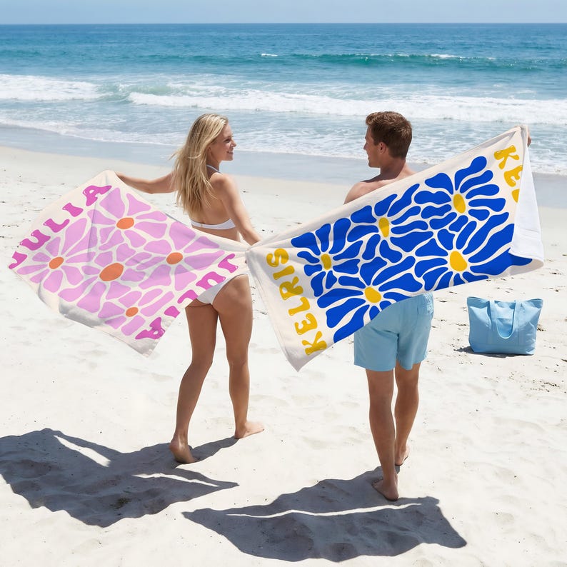 May include: Two rectangular beach towels featuring large daisy patterns. One towel is white with pink flowers and the name JULIA, while the other is white with blue flowers and the name KELRIS. Both towels have yellow flower centers.