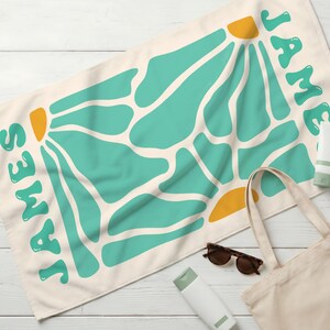 May include: A rectangular cream-colored towel featuring a repeating abstract pattern of teal organic shapes and yellow semi-circles. The name JAMES is printed in large, rounded teal letters along the left and right edges of the fabric.