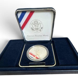 May include: A United States Mint proof coin encased in a clear protective capsule, housed within a dark blue velvet-lined display box. The interior lid features the United States Mint seal, text, and a red, white, and blue ribbon accent.