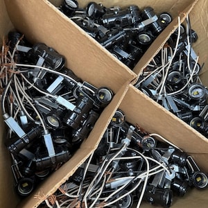 May include: A cardboard box filled with numerous black light bulb sockets with attached wires. The sockets have a cylindrical shape and are made of a glossy black material. The wires are white and black, with exposed copper ends.