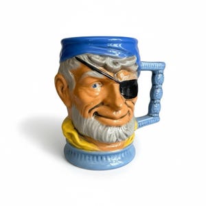 May include: A ceramic mug shaped like a pirate's head, featuring a blue bandana, eye patch, and a handle. The mug has a detailed, hand-painted design with orange, gray, and yellow accents. The mug is a unique decorative item.