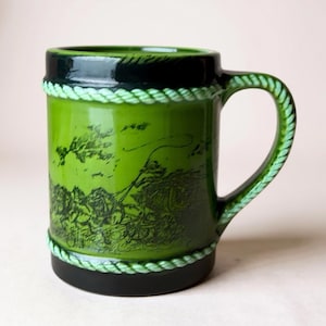 Op de afbeelding: Groene keramische mok met een zwarte rand en basis, met een touwachtig ontwerp rond de boven- en onderkant. De mok heeft een handvat en een gedetailleerde zwarte illustratie van een landschap. De mok is ongeveer 10 cm hoog.