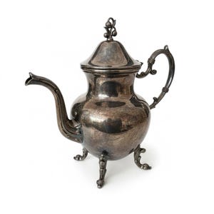 May include: A vintage silver teapot with a curved spout, ornate handle, and lid. The teapot has a bulbous body and stands on three decorative feet. The lid is topped with a small finial. The teapot is highly reflective.