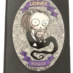 May include: A black hardcover book with a cartoon illustration on the cover. The image shows a character with long hair and skull details, holding a cat. The title "LENORE" is above, and "NOOGIES" below, on purple banners.