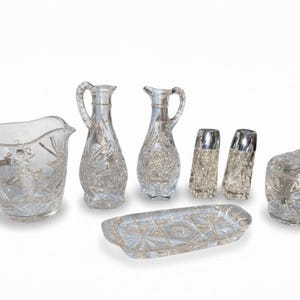 May include: A clear, cut-glass serving set featuring a creamer, two small pitchers, two salt and pepper shakers, a covered sugar bowl, and a rectangular tray. The set has a starburst pattern and is ideal for serving beverages and condiments.
