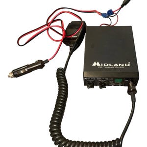 May include: Black Midland CB transceiver with a coiled microphone cord and a cigarette lighter adapter. The unit has a black exterior with the brand name and model number printed on the front. Red and black wires are visible.