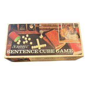 May include: A vintage Scrabble Sentence Cube Game box. The box has a dark background with the game's title and logo in white and red. The box includes a red cup, dice, and a picture of people playing the game.