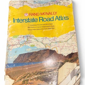 Może przedstawiać: Zabytkowy atlas drogowy Rand McNally z żółtą okładką, przedstawiający mapę stanu Waszyngton, fotografię krajobrazu przybrzeżnego oraz tekst "Completely Revised and Up to Date" i "800 Points of Interest Organized by State."
