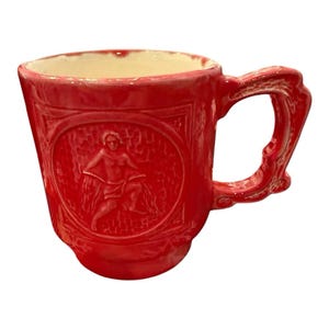 May include: A vibrant red ceramic mug with an ornate handle and a detailed relief design. The relief features a classical figure within an oval frame. The interior of the mug is a cream color, contrasting with the exterior.
