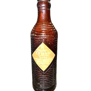 Puede incluir: Una botella de refresco de vidrio marrón vintage con una superficie texturizada y acanalada y una etiqueta en forma de diamante. La etiqueta presenta el texto Orange Crush Soda y 7 FL. OZS. La botella tiene un cuello estrecho y un borde redondeado.