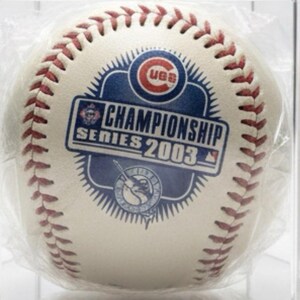 Puede incluir: Una pelota de béisbol con costuras rojas y un diseño impreso que conmemora la Serie de Campeonato de 2003. El diseño incluye el texto "CHAMPIONSHIP SERIES 2003" y logotipos de equipos. La pelota está dentro de una vitrina transparente.