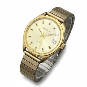 May include: A gold-toned Benrus automatic wristwatch with a cream-colored face and a date display at the 3 o'clock position. The watch features a gold-toned bezel and a stretch band. The watch face has the text "BENRUS" and "25 JEWELS AUTOMATIC".