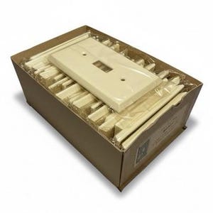 Op de afbeelding: Een kartonnen doos gevuld met crèmekleurige elektrische schakelplaten en afdekkingen. De doos bevat meerdere platen en afdekkingen, sommige individueel verpakt in doorzichtig plastic. De doos is open en toont de inhoud.