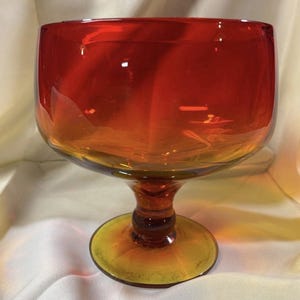 May include: A glass bowl with a wide, rounded top that transitions from a deep red at the rim to a warm amber color at the base. The bowl is supported by a thick, short pedestal stem and a circular foot, all crafted from smooth, translucent glass.