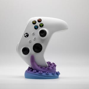 Puede incluir: Un mando blanco de Xbox se muestra sobre un soporte con un diseño inspirado en un pulpo. El soporte presenta un degradado de azul y morado, con los botones del mando en varios colores. El mando es el foco principal, mostrando su diseño elegante.