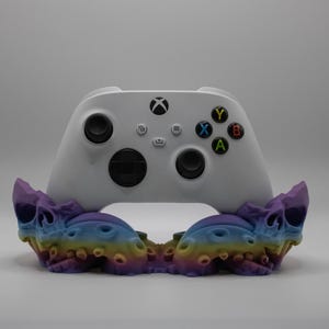 Puede incluir: Un mando blanco de Xbox descansa sobre un soporte colorido con forma de calavera. El soporte presenta un degradado de tonos morados, azules, amarillos y naranjas. El mando tiene botones negros y botones de acción de colores.