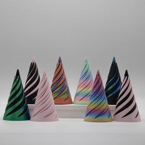 May include: A group of cone-shaped decorative objects in shades of green, black, pink, and rainbow. Each cone has a spiral pattern. The objects are arranged on a white surface. The design is eye-catching and colourful.