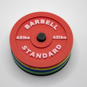 May include: A red barbell weight plate with white lettering that reads "BARBELL STANDARD" and "45lbs". The weight plate is stacked on top of several other plates with different coloured edges, including blue, green, yellow, and black.