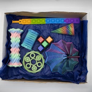 Könnte beinhalten: Eine Schachtel voller bunter 3D-gedruckter Spielzeuge. Enthält ein Regenbogen-Push-Pop-Fidget-Spielzeug, einen Kegel, ein Zahnrad, einen Fächer und andere geometrische Formen in Blau-, Grün-, Rosa- und Lilatönen. Die Schachtel ist mit blauem Seidenpapier ausgekleidet.