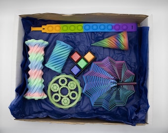May include: A box filled with colorful 3D-printed toys. Includes a rainbow push-pop fidget toy, a cone, a gear, a fan, and other geometric shapes in shades of blue, green, pink, and purple. The box is lined with blue tissue paper.