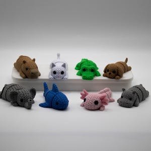 Op de afbeelding: Een verzameling handgemaakte gehaakte dierenfiguren, waaronder een bruin nijlpaard, een witte kat, een groene kikker, een bruine hond, een grijze olifant, een blauwe haai, een roze axolotl en een grijs nijlpaard. De figuren zijn gerangschikt op een wit oppervlak.