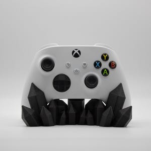 Op de afbeelding: Een witte Xbox-controller met zwarte accenten, rustend op een zwarte, kristalachtige basis. De controller heeft gekleurde knoppen en het Xbox-logo. Een modern gaming accessoire.