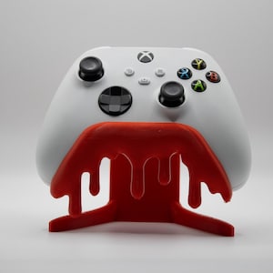 以下が含まれることがあります： 白いXboxコントローラーが鮮やかな赤いコントローラーのスタンドに置かれています。スタンドは、滴るような効果を持つユニークなデザインです。コントローラーには、黒いサムスティックとカラフルなアクションボタンが付いています。
