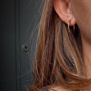 May include: A simple gold hoop earring with a smooth, polished finish worn in the earlobe. The earring features a circular design with a thin, rounded band that creates a classic look. The metal has a warm, reflective yellow gold tone.