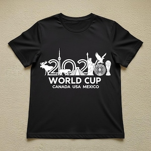 May include: Black t-shirt with a white graphic design. The design includes the text "2026 WORLD CUP CANADA USA MEXICO" along with illustrations of a moose, a panther, the CN Tower, the Statue of Liberty, an eagle, and a trophy.