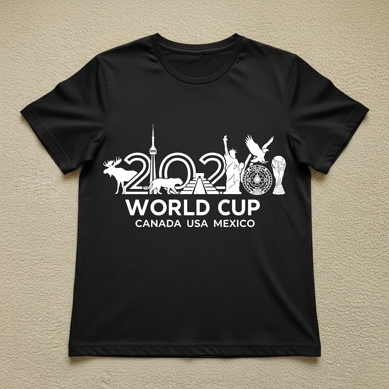 May include: Black t-shirt with a white graphic design. The design includes the text "2026 WORLD CUP CANADA USA MEXICO" along with illustrations of a moose, a panther, the CN Tower, the Statue of Liberty, an eagle, and a trophy.