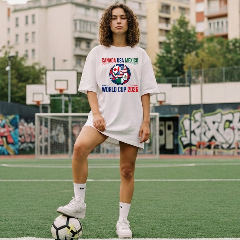 May include: White t-shirt with the text "CANADA USA MEXICO WORLD CUP 2026" in red, green, and blue. The shirt features a soccer ball design with the flags of Canada, the United States, and Mexico. The person is standing on a soccer ball.