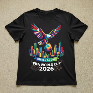 May include: Black t-shirt featuring a colorful eagle with outstretched wings perched atop a soccer ball. The design includes a cityscape and the text "UNITED AS ONE FIFA WORLD CUP 2026" with fireworks.