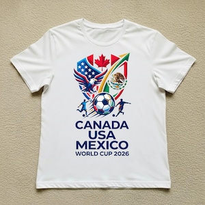 May include: White t-shirt featuring a colorful graphic design for the 2026 World Cup. The design includes the flags of Canada, the United States, and Mexico, along with a soccer ball and the text "CANADA USA MEXICO WORLD CUP 2026".