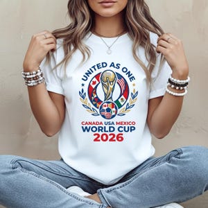 May include: White t-shirt featuring a colorful graphic for the 2026 World Cup, with the text "UNITED AS ONE" and "CANADA USA MEXICO WORLD CUP 2026". The design includes a trophy, a soccer ball, and flags.
