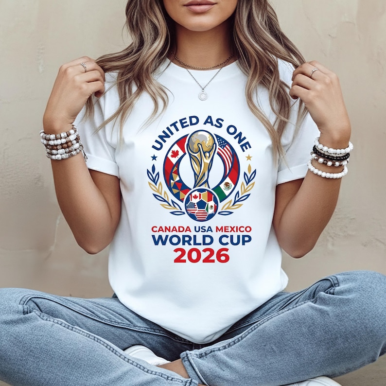 May include: White t-shirt featuring a colorful graphic for the 2026 World Cup, with the text "UNITED AS ONE" and "CANADA USA MEXICO WORLD CUP 2026". The design includes a trophy, a soccer ball, and flags.